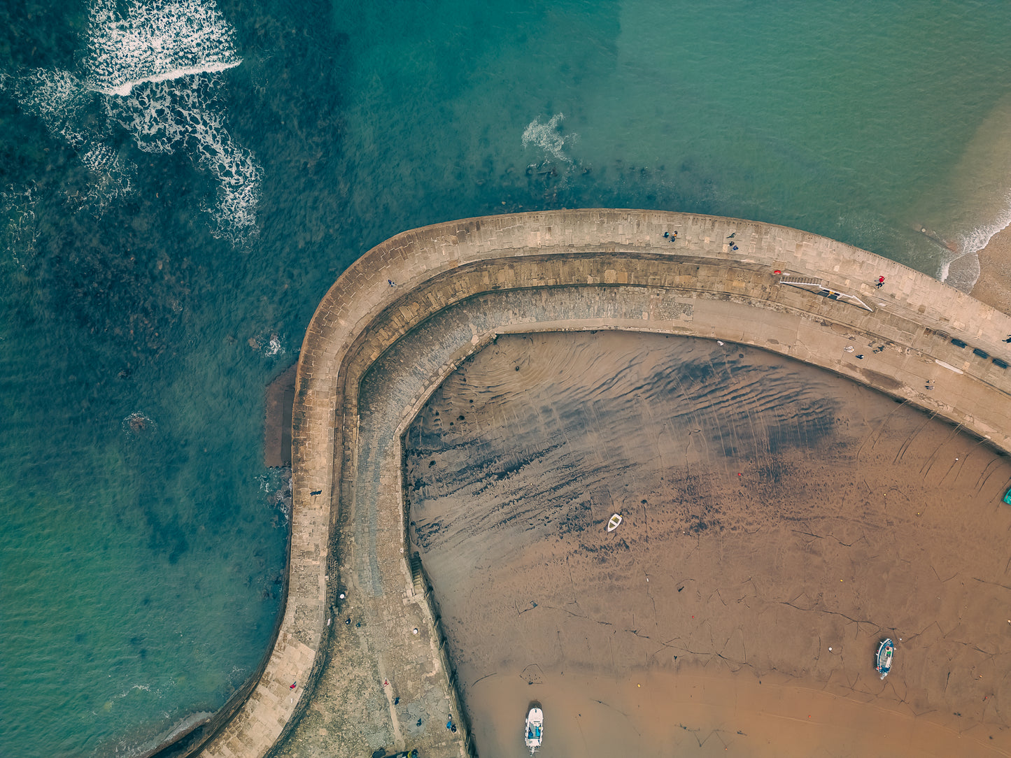 Aerial view of the Cobb at Lyme Regis showing the curved stone harbour wall between turquoise sea and textured low-tide sand, with small boats resting on the shore.